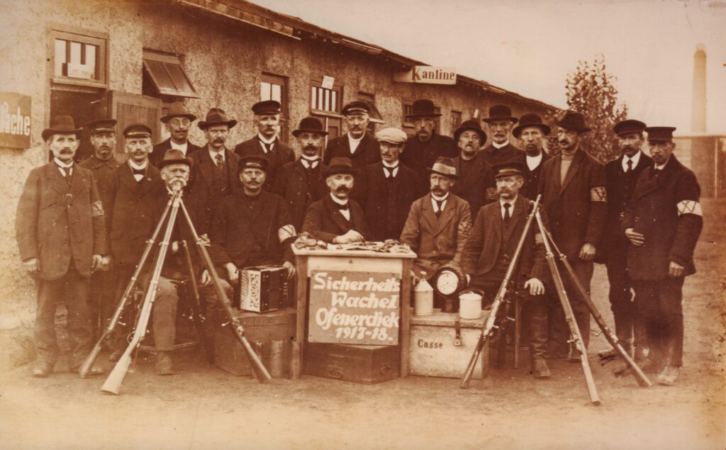 Eine Gruppe von Männern in Anzügen posiert für ein Foto, vor ihnen sind Gewehre aufgestellt, in der Mitte ein Schild mit Beschriftung, im Hintergrund ein Fabrikgelände, © Stadtmuseum Oldenburg