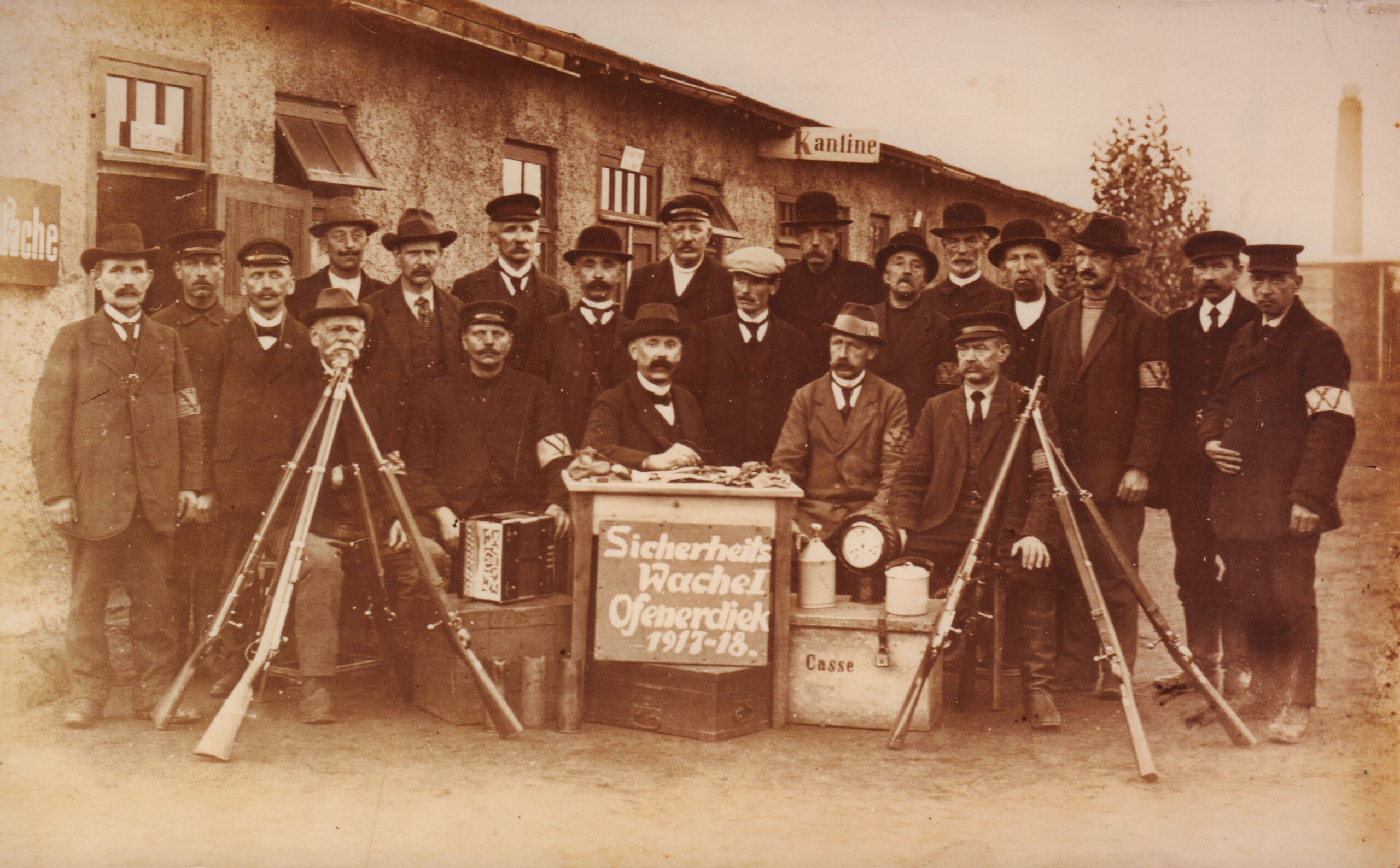 Eine Gruppe von Männern in Anzügen posiert für ein Foto, vor ihnen sind Gewehre aufgestellt, in der Mitte ein Schild mit Beschriftung, im Hintergrund ein Fabrikgelände, © Stadtmuseum Oldenburg