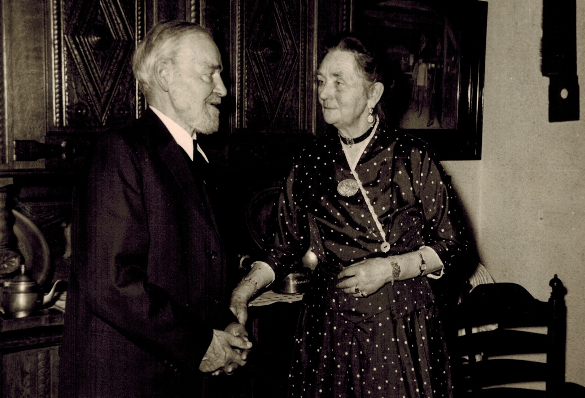 Bernhard Winter und seine Ehefrau Martha um 1955, © Stadtmuseum Oldenburg Zwei ältere Menschen unterhalten sich, Er trägt einen Anzug, sie ein Kleid mit Punkmuster, sie stehen in einem Raum mit Holzmöbeln, © Stadtmuseum Oldenburg