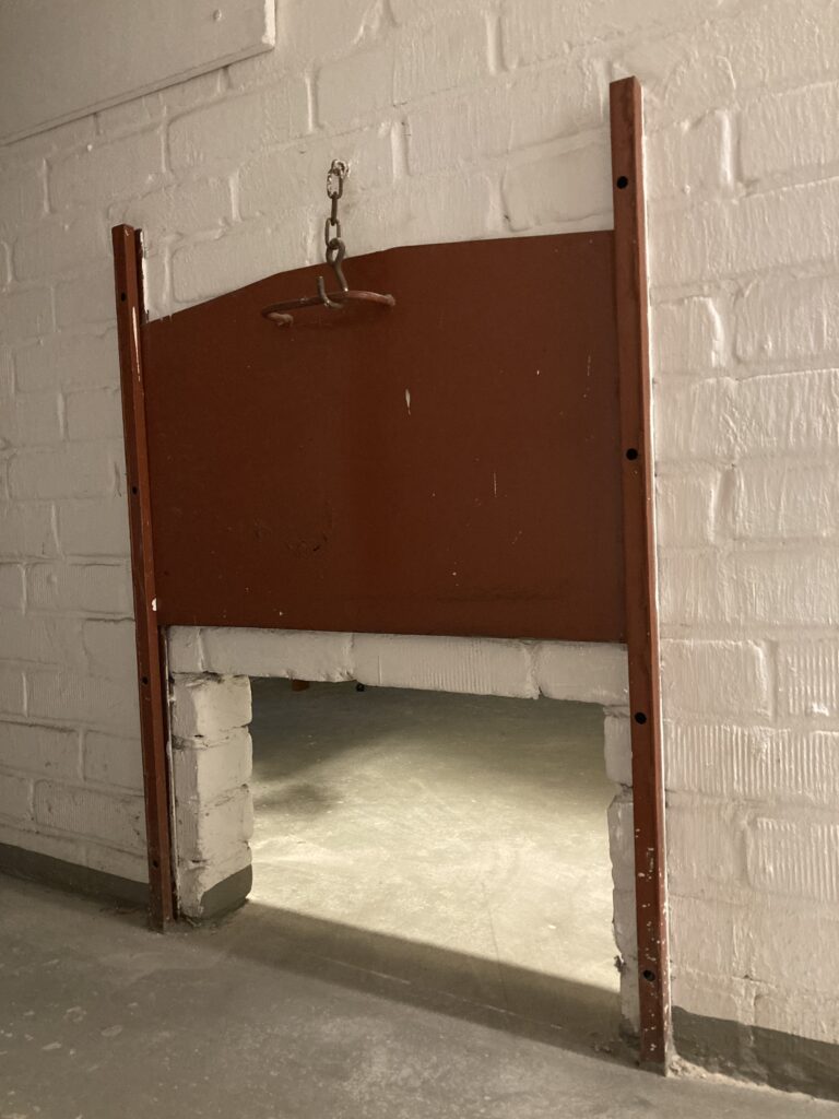 Die frühere Nutzung des Kellers als Kohlenkeller ist im heutigen Haus noch deutlich durch die metallene Hebeklappe zu erkennen, © Privatbesitz Weiß verputzte Backsteinwand mit einer roten Metallklappe, niedrige Öffnung zum Nachbarraum, © Privatbesitz