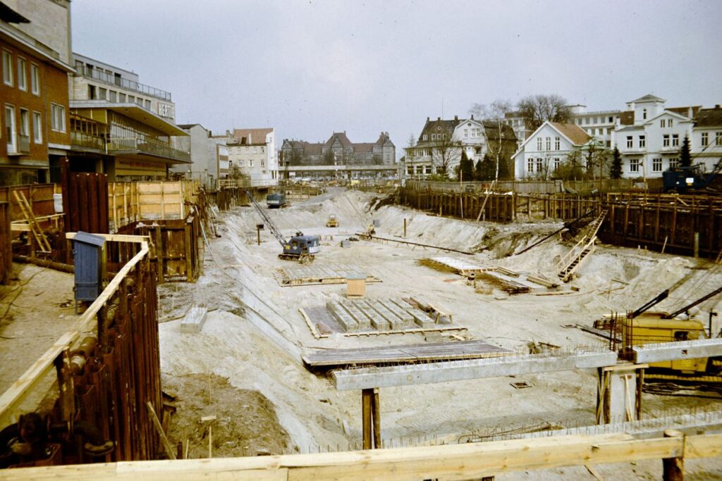 Baustelle eines großen Straßenbaus, Baugrube mit Baustellenfahrzeugen und Baumaterial, links und rechts davon Häuserzeilen, © W. Meyer/Stadtmuseum Oldenburg