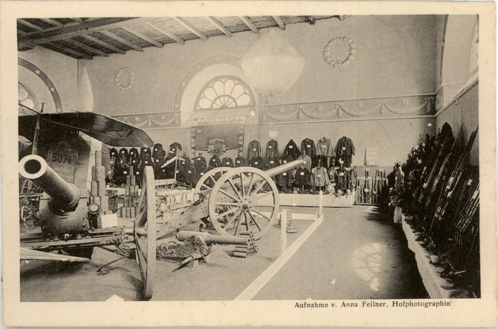 Kriegsgeräte, darunter Kanonen und Gewehre in einer Halle, an der hinteren Wand hängen Uniformen, © Stadtmuseum Oldenburg.