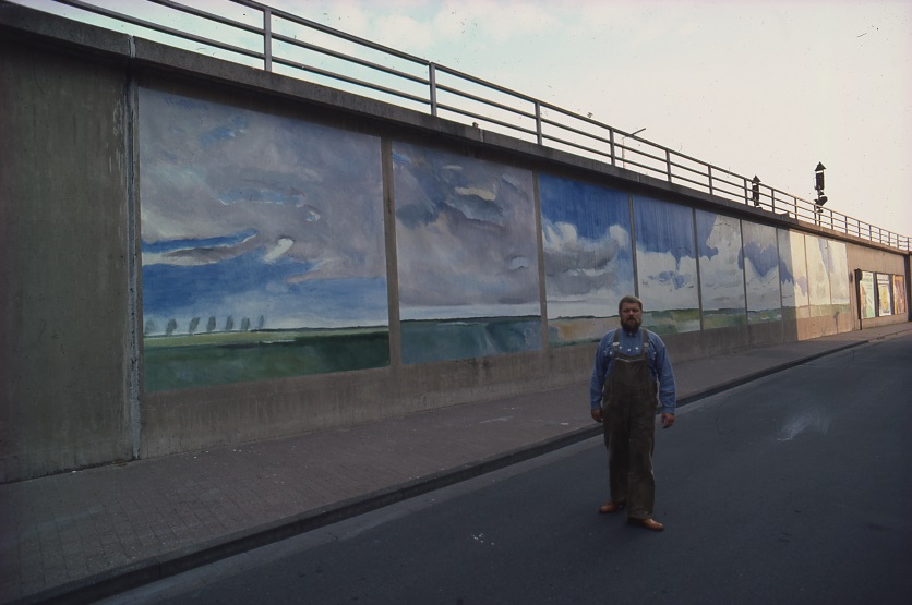 Abb.3 Mehrere Banner mit Landschaftsmalerei hängen an einer Mauer, ein Mann steht auf der Straße davor, © Gerresheim/Stadtmuseum Oldenburg