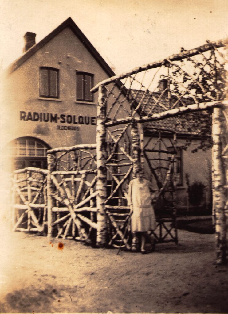 Abb.5 BA 18483 1930 Slg Schäfer Eine Person steht an einem Holztor mit strahlenförmigem Muster, dahinter ein Haus mit Aufschrift "Radium-Solquelle", © Stadtmuseum Oldenburg