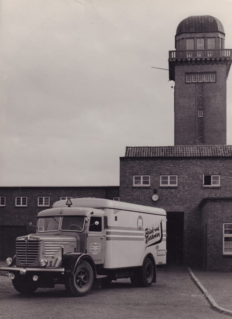 04 BA 32495 GEG Fleiwa Auslieferungsfahrzeug Um 1930 Firmenarchiv Ein Auslieferungsfahrzeug steht im Vordergrund eines Klinkergebäudes. Im Hintergrund ist ein Wasserturm zu sehen.