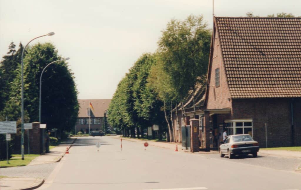 Die Einfahrt des Fliegerhorstes im Jahr 1998 zeigt sich wenige Jahre vor der Schließung, als eine lange Straße an deren Rand eine Pforte zu sehen ist. Die Straße ist mit Pylonen und Beschilderung vor Durchfahrt geschützt.