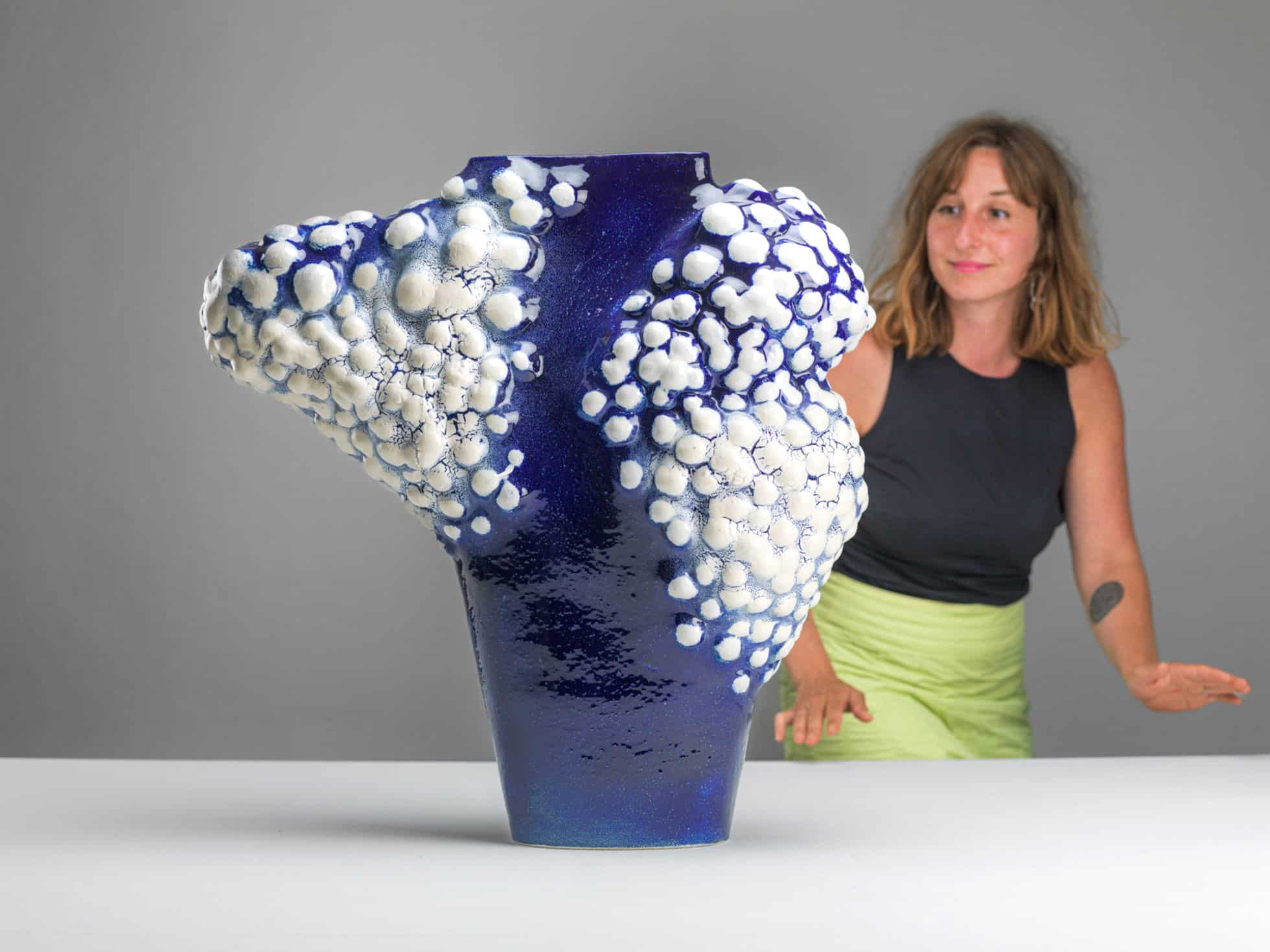 Maya Fenderl, Blue Bird, Weißer Ton, Glasur, 2024 Foto Alexander Burzik Skulptur aus blau glasiertem Ton mit weißen, kugeligen Erhebungen auf einem Tisch vor der Künstlerin in schwarzem Oberteil und grünem Rock