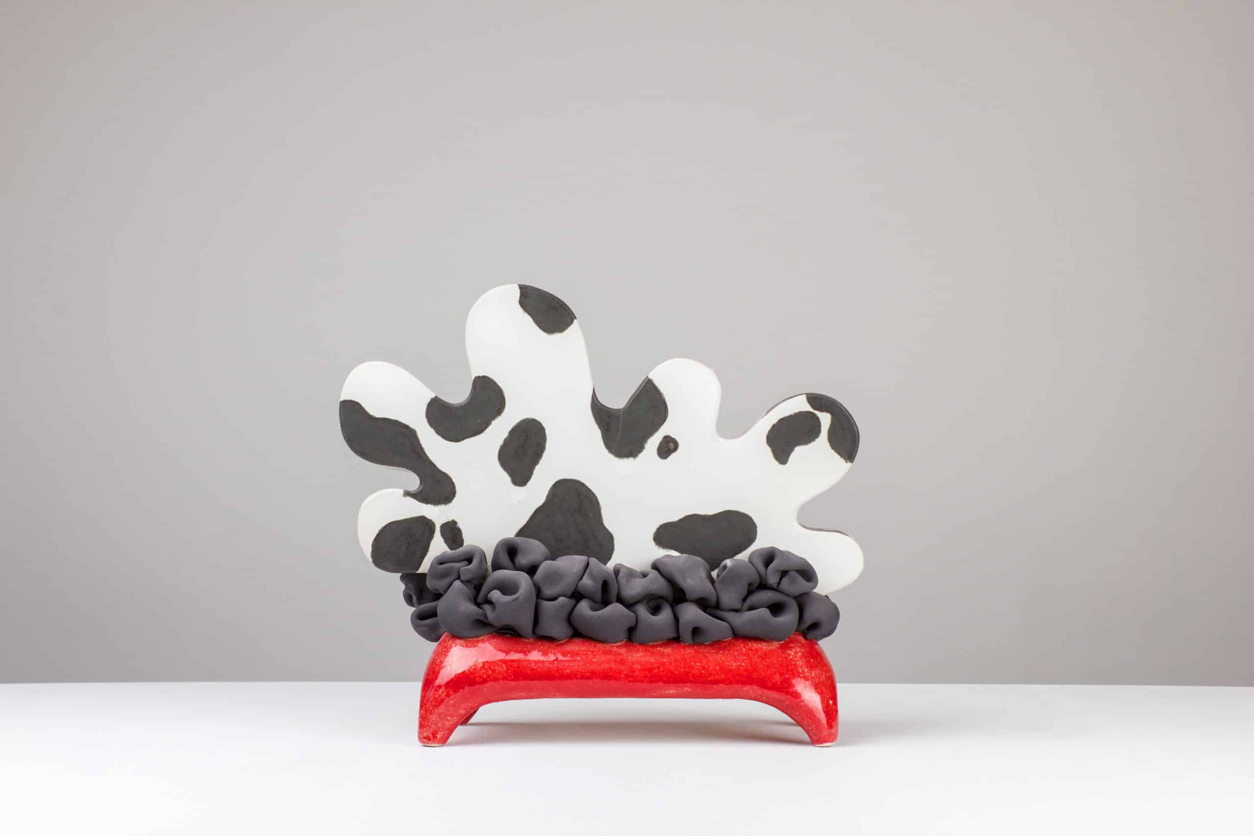 Resting Cow, Weißer Ton, Engobe, Glasur, 2026 © Maya Fenderl (2) Keramische Skulptur einer liegenden Kuh mit schwarz-weißer Fleckenzeichnung, grauem, knautschigem Unterbau und rotem Sockel