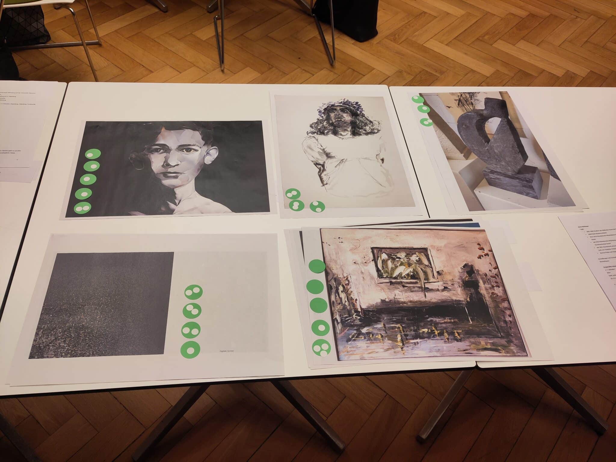 Auswahl Jury Stadtmuseum Oldenburg Sechs Kunstwerke auf einem Tisch mit grünen Bewertungsaufklebern darunter, darunter Porträt, Skulptur und abstrakte Malerei