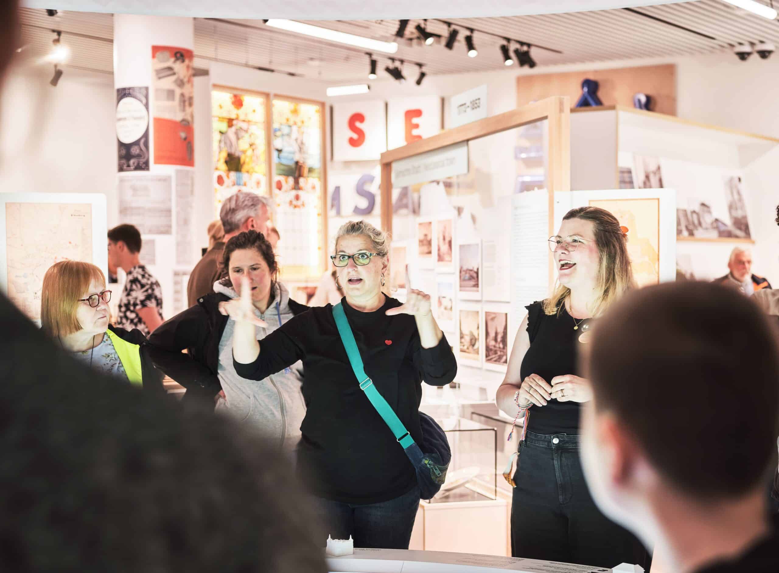 CS20260607 SMO Opening 5310 Gruppe von Menschen in einem hellen Ausstellungsraum, eine Frau mit Brille und grünem Schulterriemen gestikuliert lebhaft