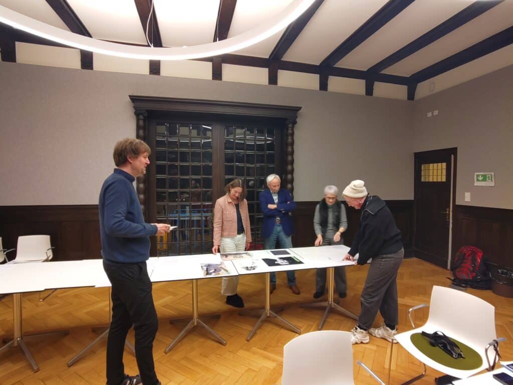 Jurymitglieder Juryankauf Stadtmuseum Oldenburg Fünf Personen stehen um einen Tisch in einem Raum mit Holzboden und sichtbaren Deckenbalken, auf dem Tisch liegen Papiere und Fotos