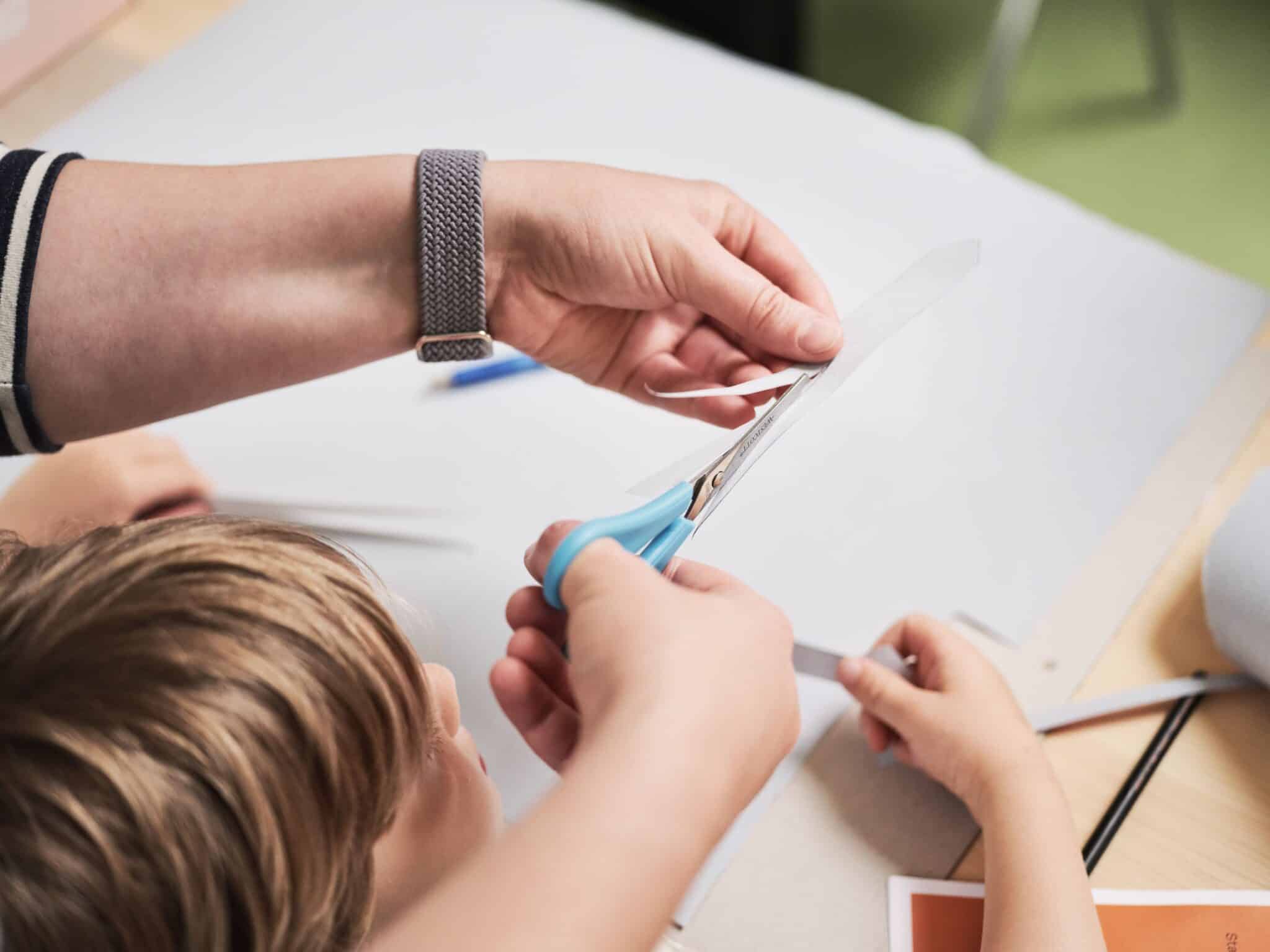 Kreativangebot Foto Caspar Sessler Hände schneiden gemeinsam mit einer Schere ein weißes Blatt Papier an einem Tisch