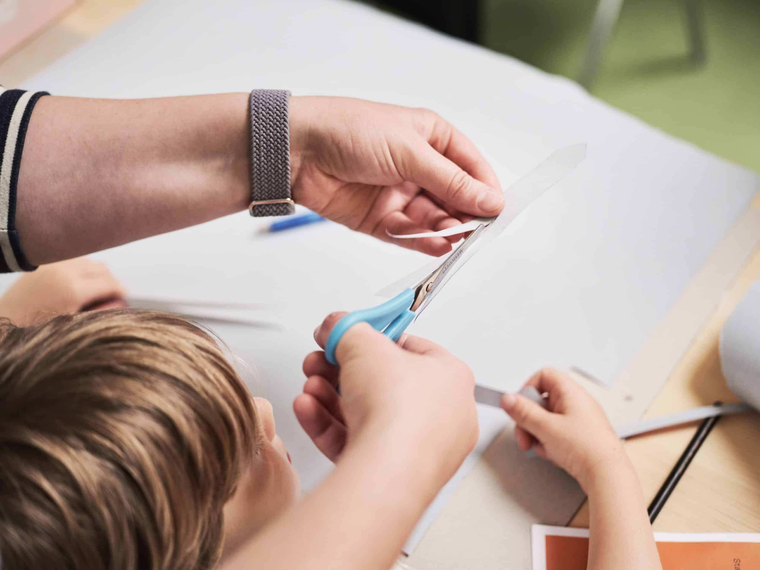 Hände schneiden gemeinsam mit einer Schere ein weißes Blatt Papier an einem Tisch